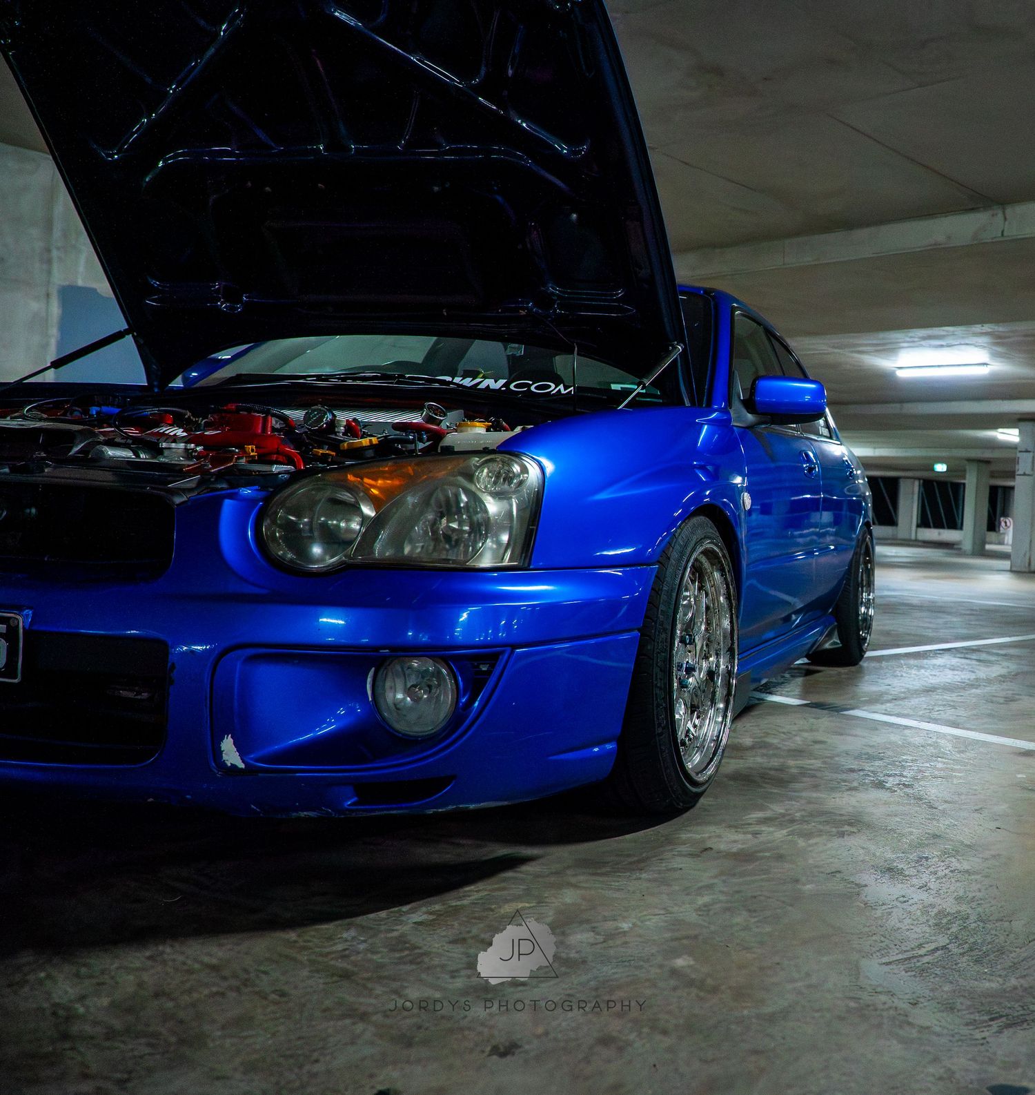 A bright blue modified sports car with aftermarket wheels parked in a dimly lit parking garage.