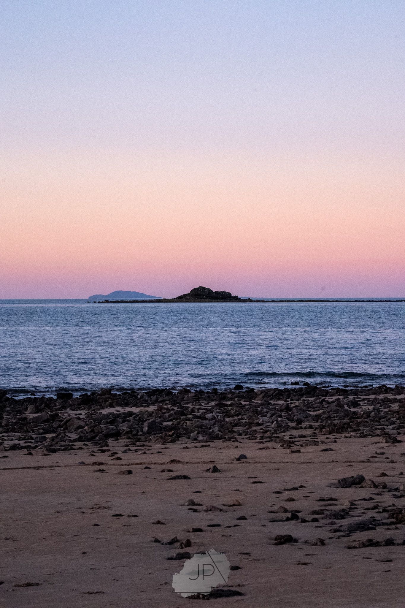 Sunset over small islands creates pink and purple hues in the sky above a rocky beach shoreline during dusk.