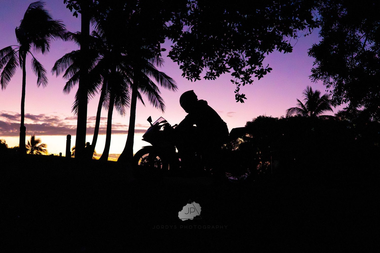 Silhouette of a motorcyclist at sunset with palm trees and purple sky creating a dramatic tropical backdrop.