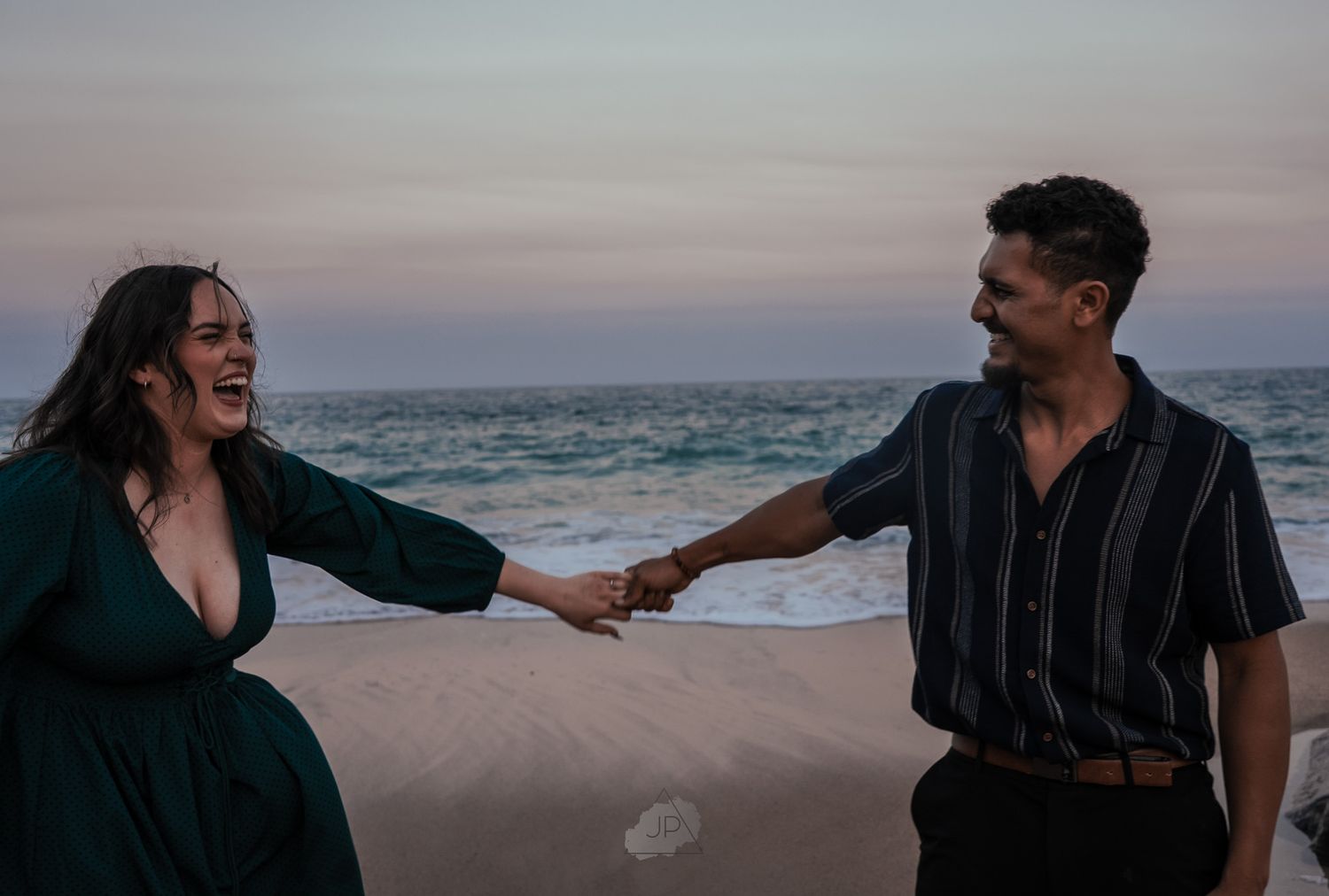 A romantic couple enjoys a sunset stroll along the beach with ocean waves and pink sky in the background.
