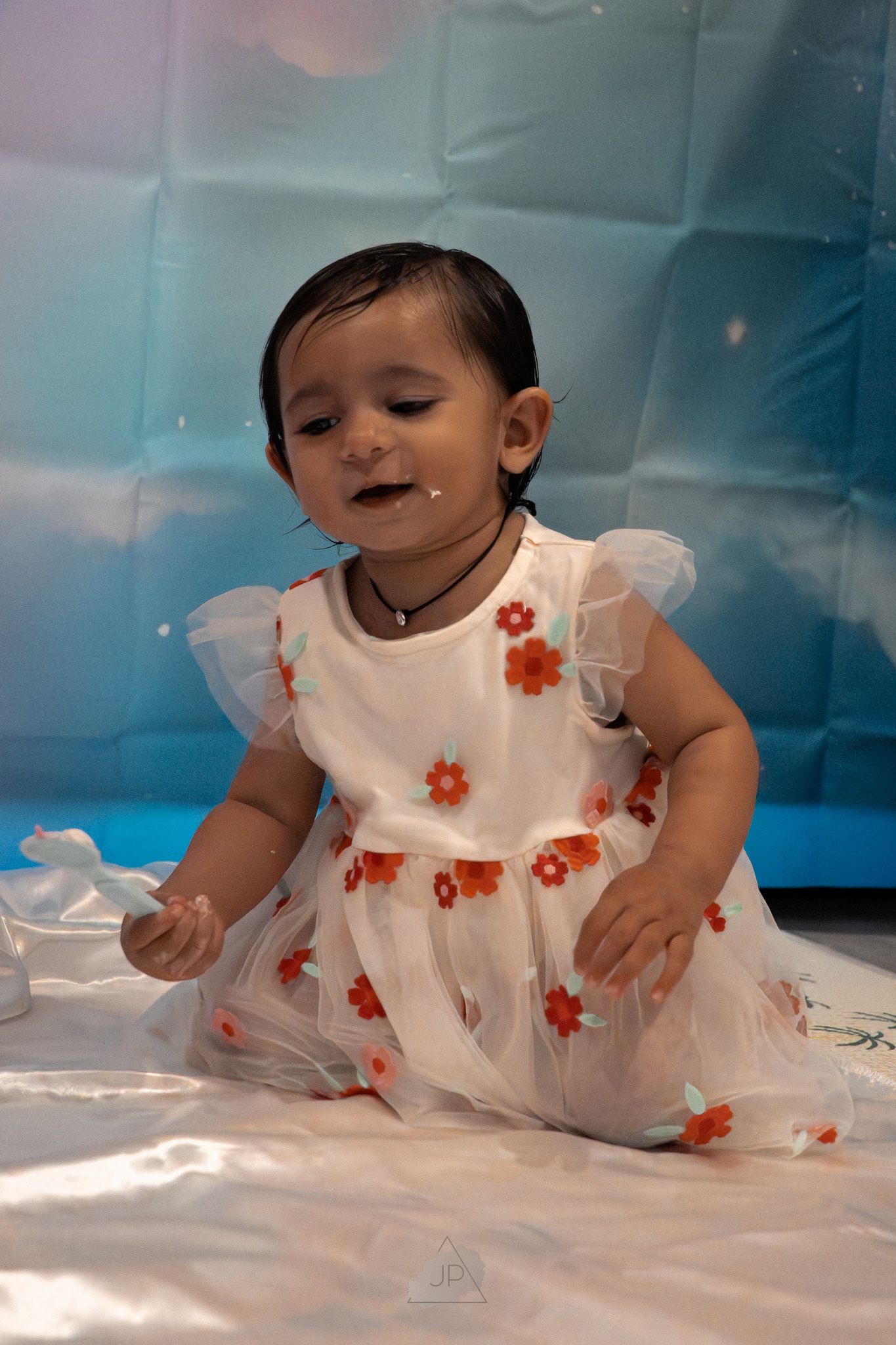 A small child in a white dress with orange flowers sits on a white surface against a blue backdrop.