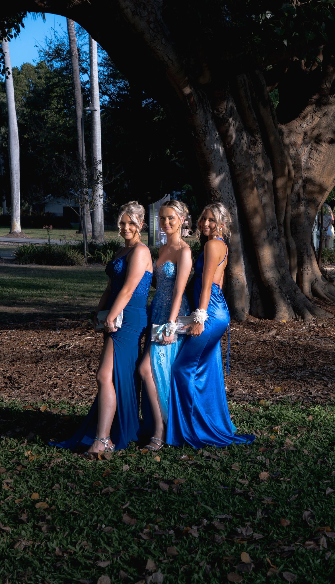 Three bridesmaids in flowing blue dresses pose together under trees in a park setting at dusk.