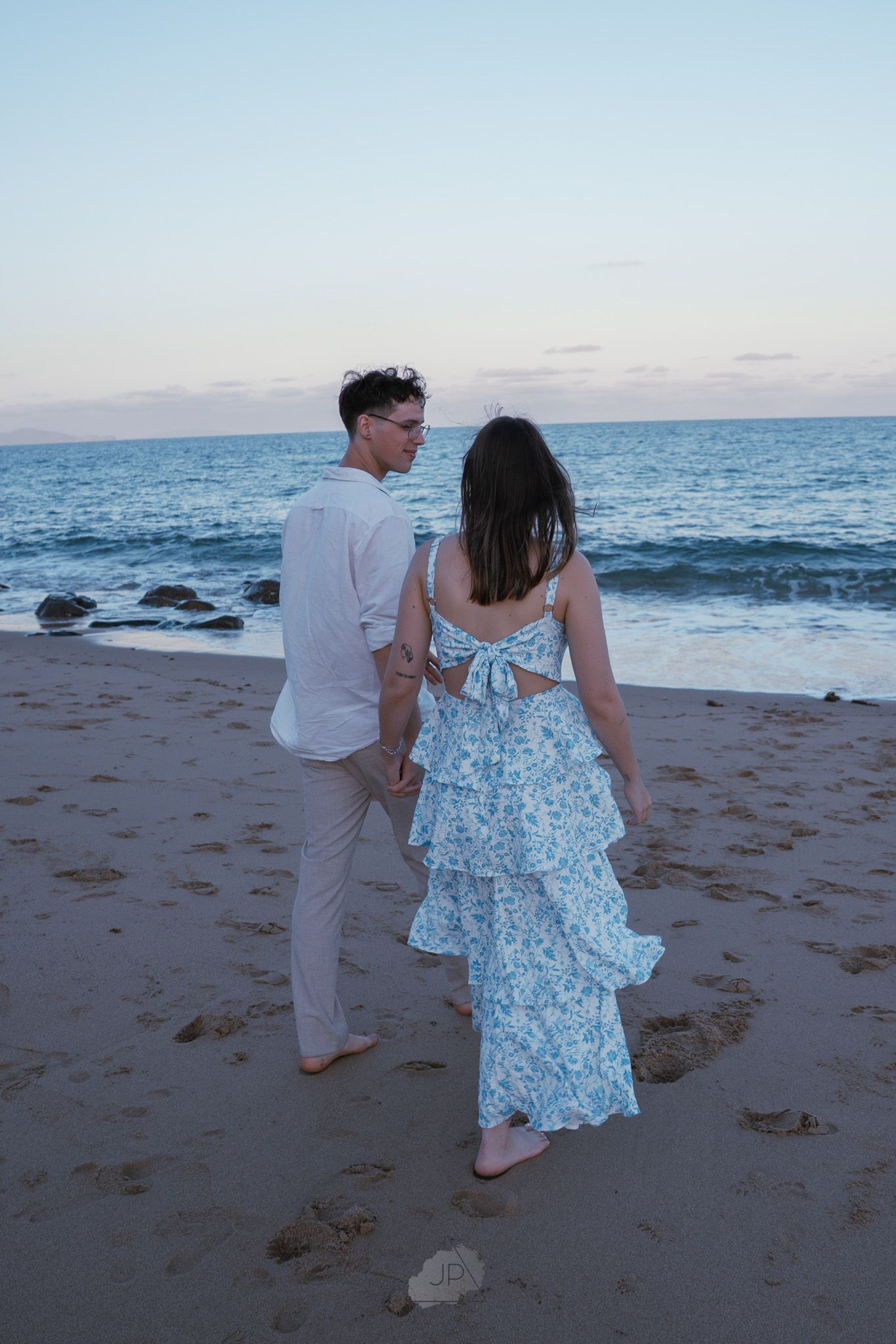 A couple walking together on a sandy beach at sunset with ocean waves and rocks in the background.