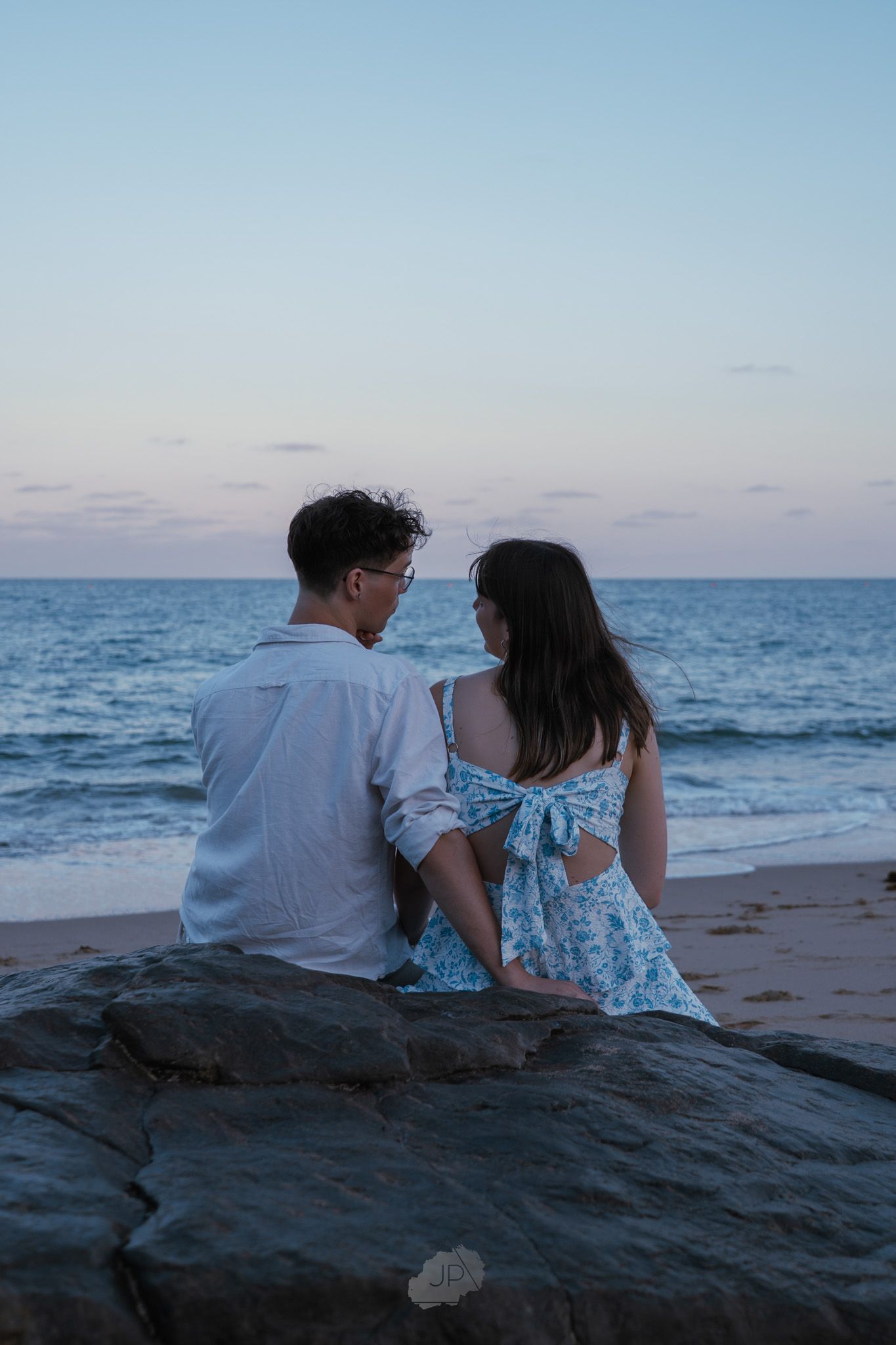 A couple sits together on dark rocks at the beach during sunset, looking out at the ocean horizon.