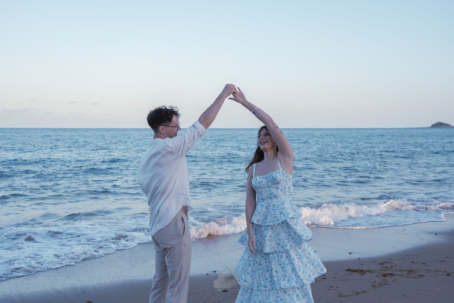 Two people in white and blue outfits hold hands while walking along a scenic beach at sunset.