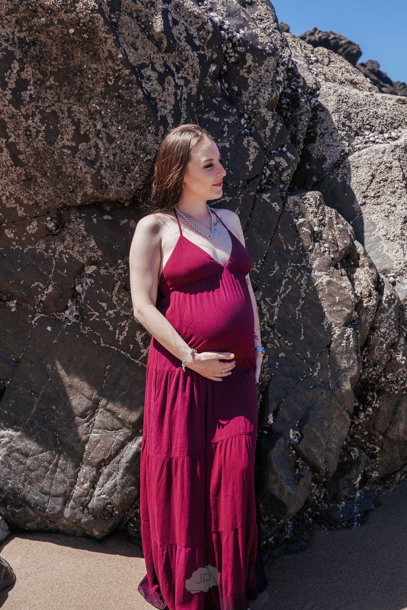 A figure in a flowing burgundy dress poses against dramatic rocky cliffs at the beach.