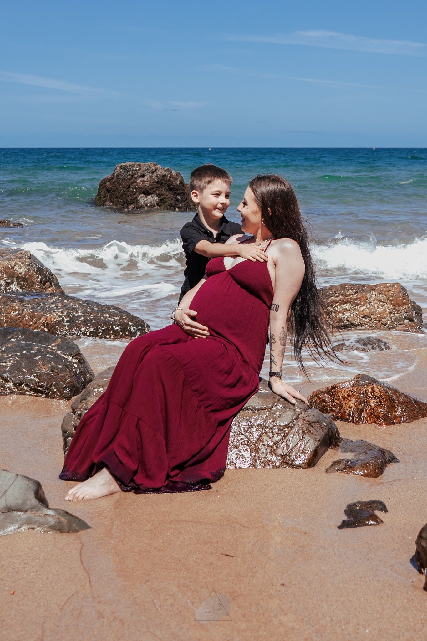 A couple in burgundy attire shares romantic moments on a sandy beach with crashing waves and rocky outcrops.