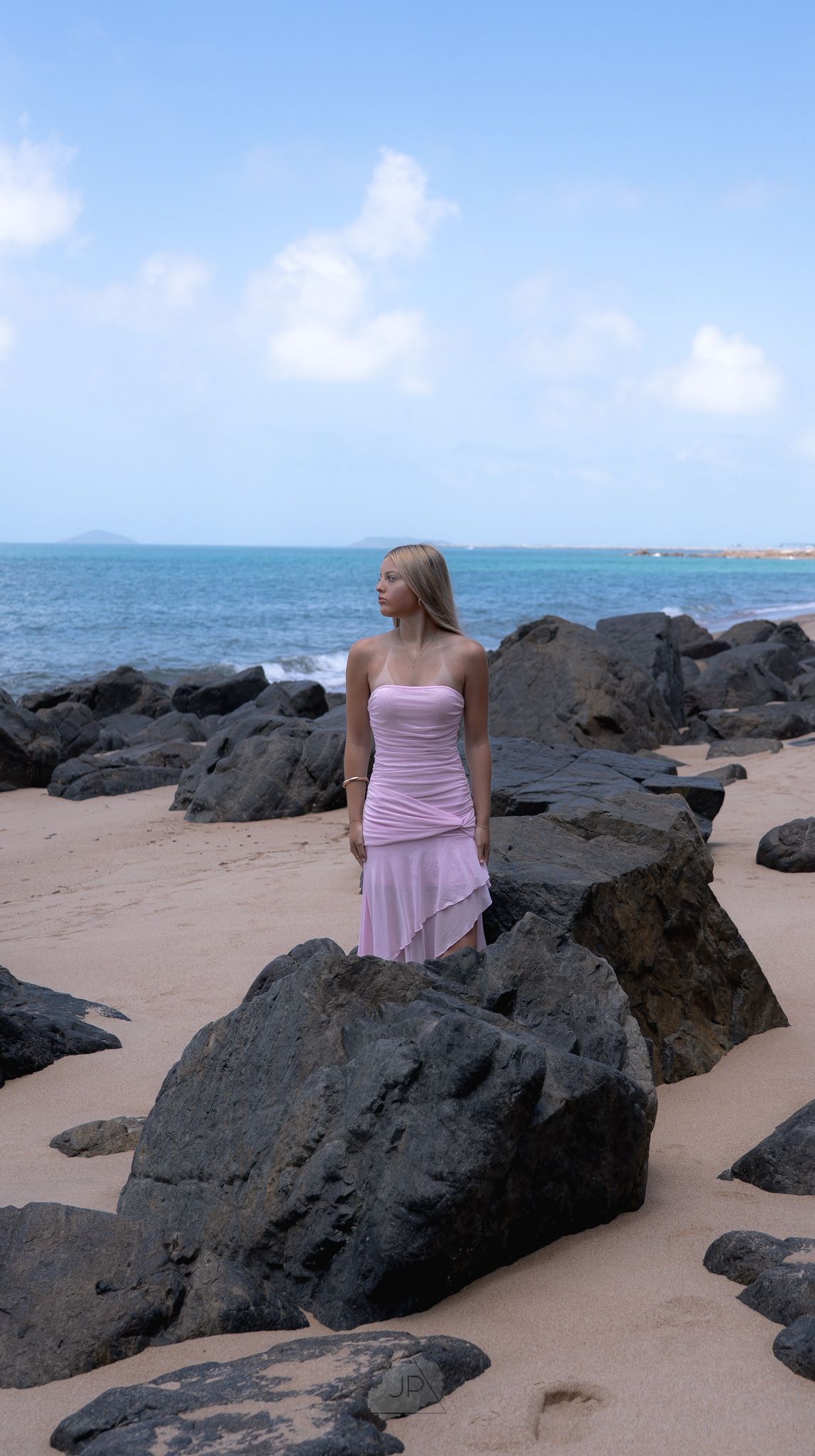 A person in a pink dress sits on black volcanic rocks at a tropical beach with turquoise water.