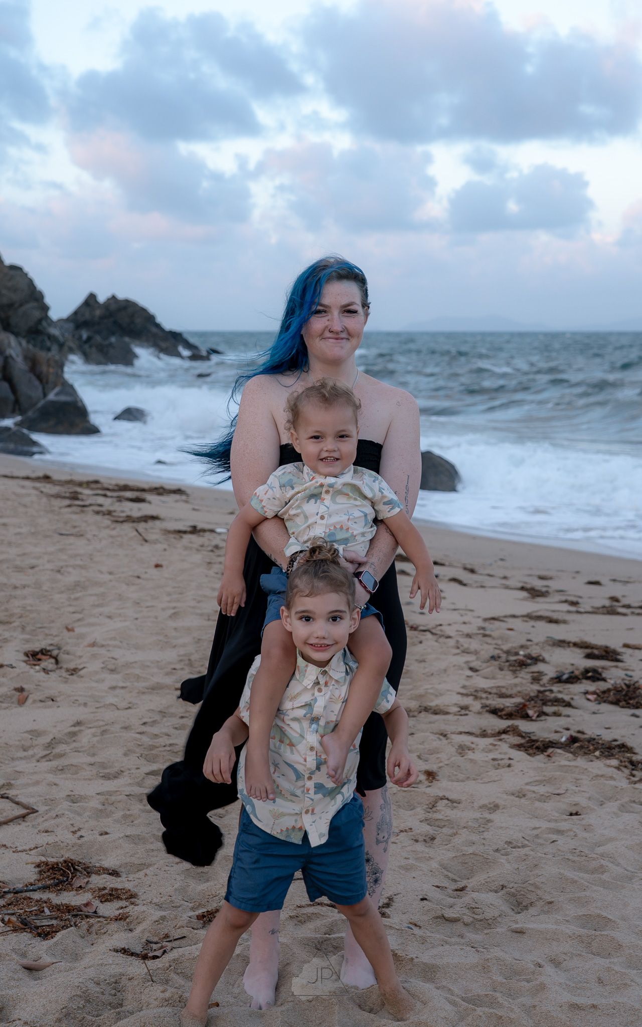 Family posing playfully on a sandy beach with ocean waves and rocky outcrops behind them.