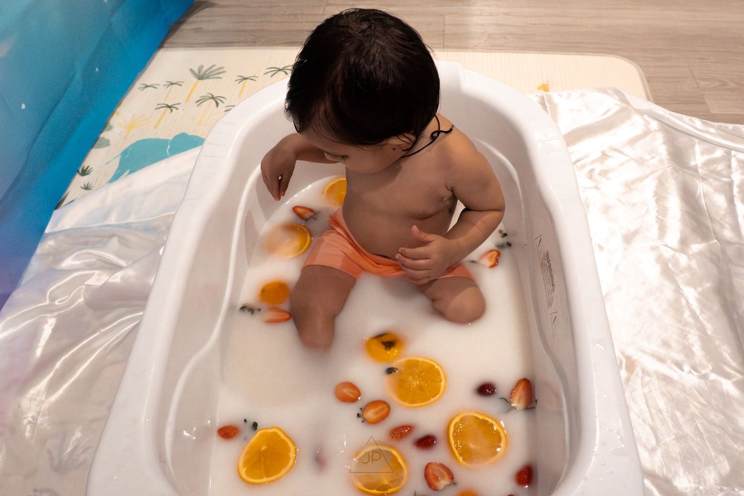 A baby sits in a white bathtub with orange citrus slices floating in the warm water during bathtime.