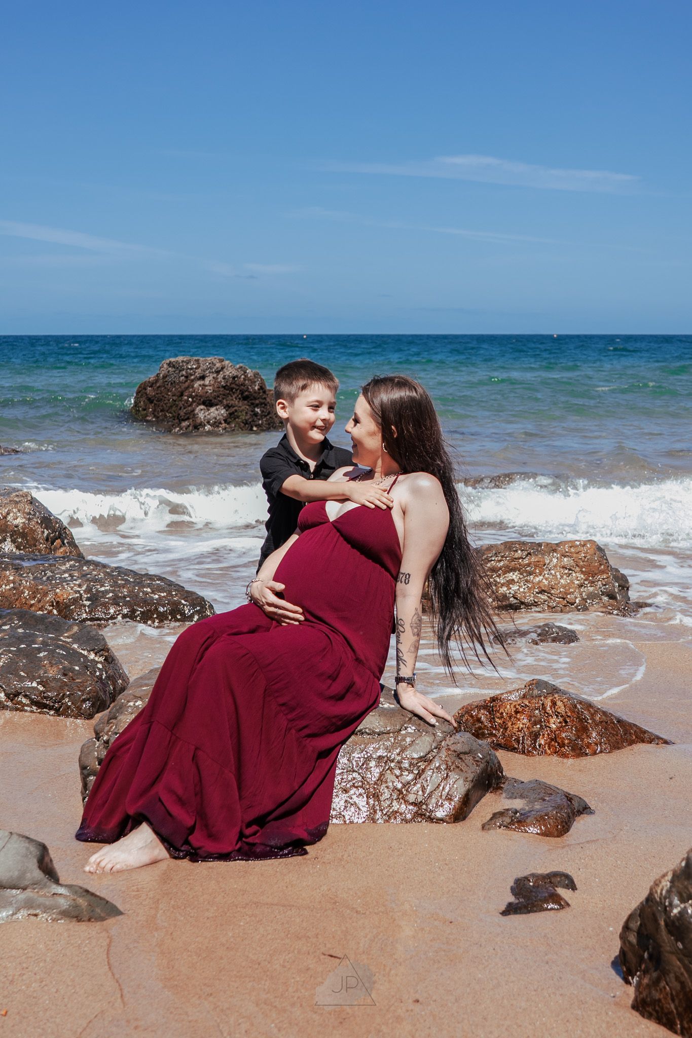 Couple in burgundy attire embracing on a rocky beach with blue ocean waves and clear sky in the background.