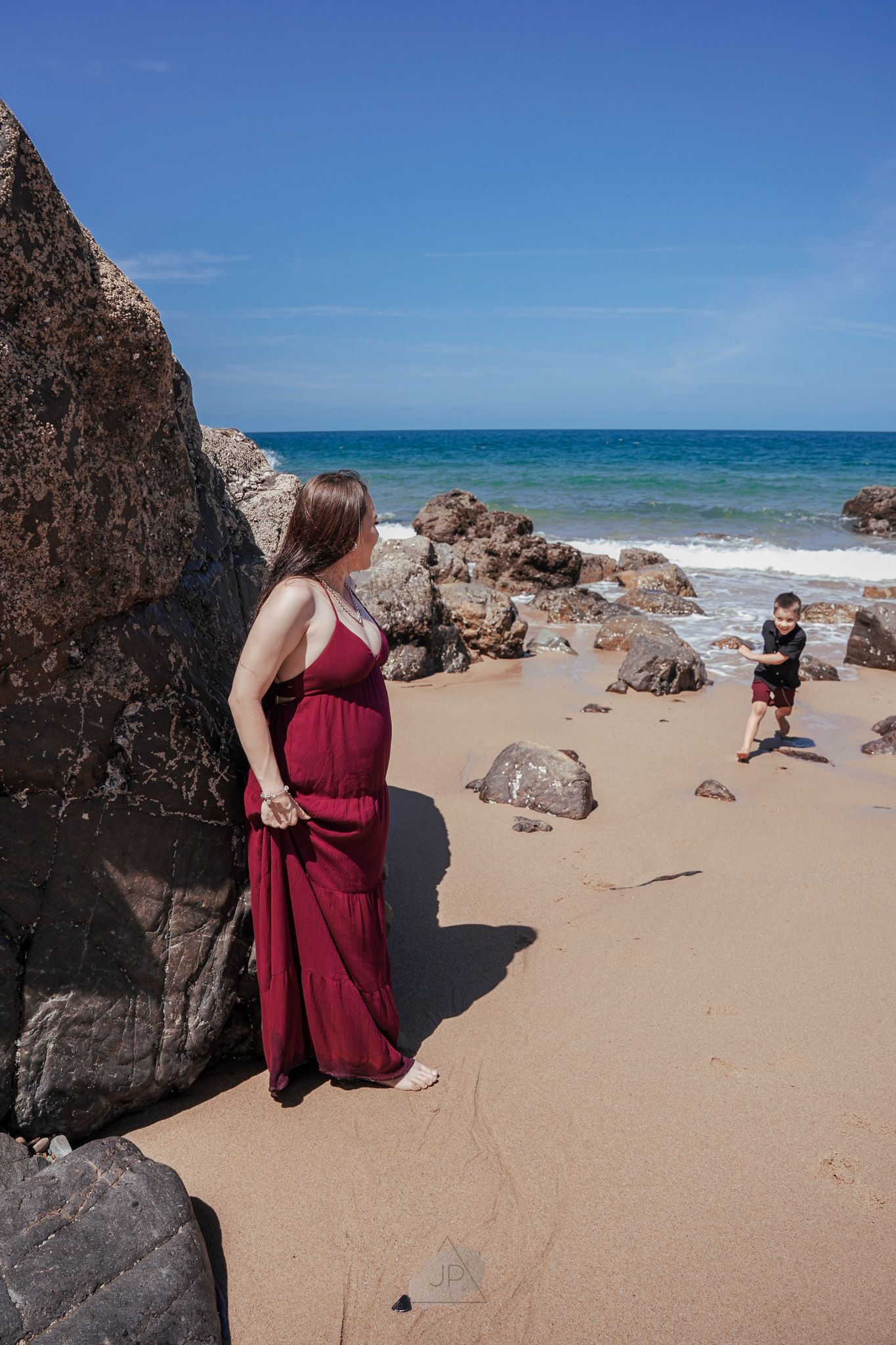 Person in burgundy dress poses on sandy beach with dramatic rocky outcrops and blue ocean under clear sky.
