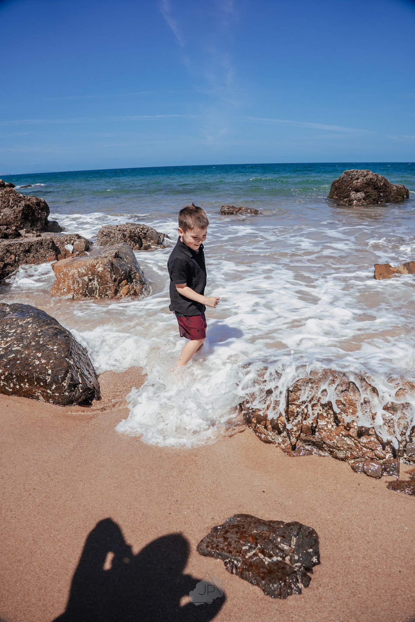 Waves crash against rocky outcrops on a sandy beach under blue skies on a sunny day.