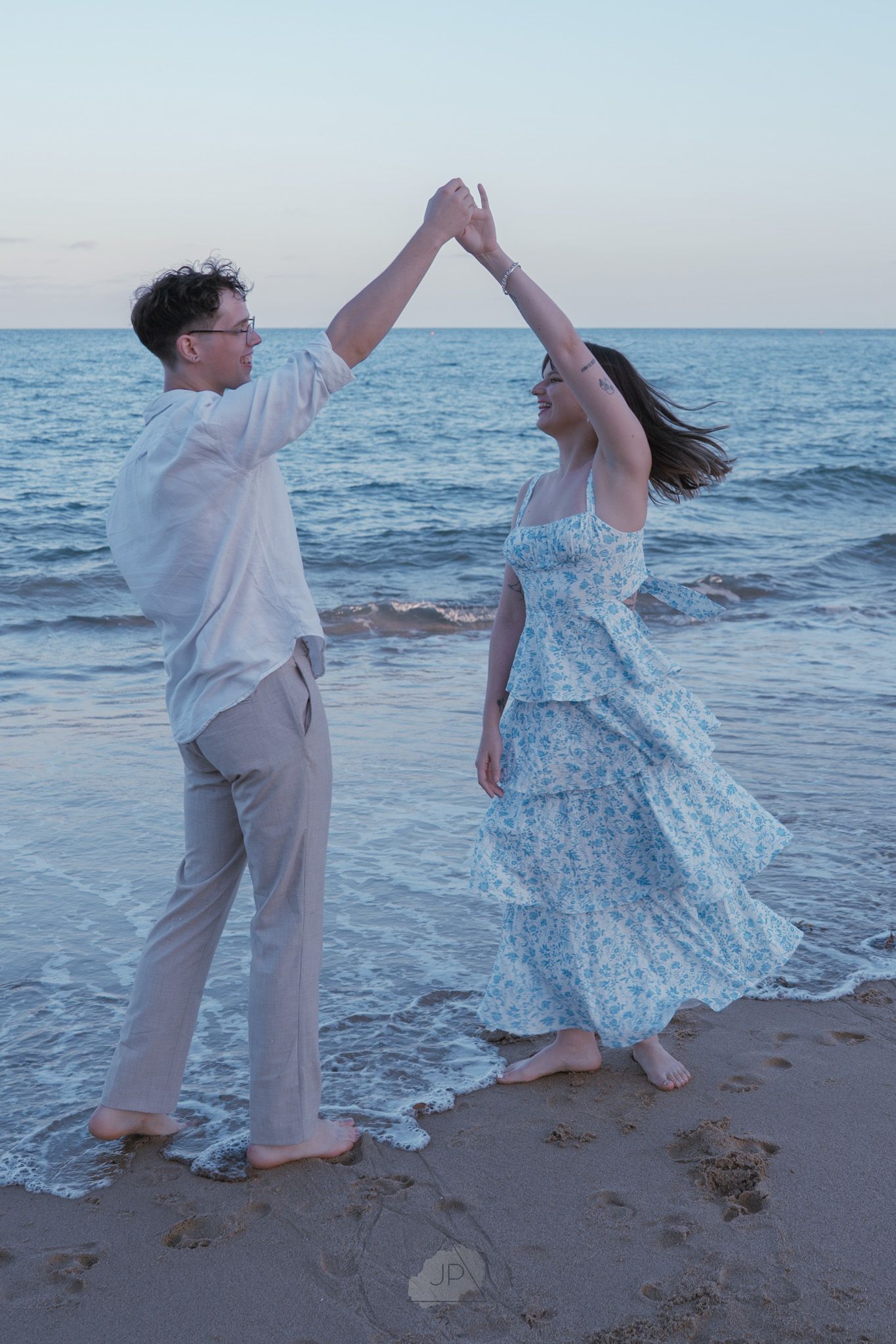 A couple dances playfully together on the beach at sunset wearing coordinated white and blue outfits.