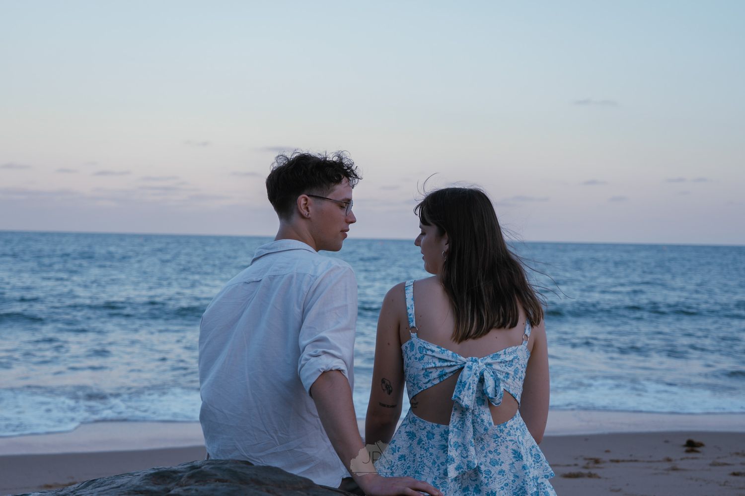 A couple sits together on the beach at dusk looking out at the ocean waves.