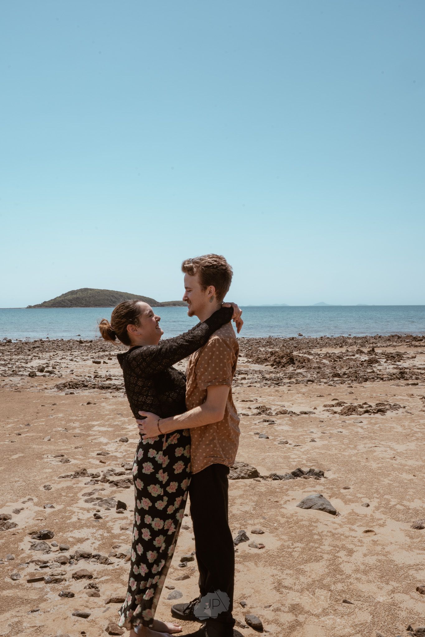 Two people embrace on a rocky beach with a distant hill visible on the horizon under a blue sky.