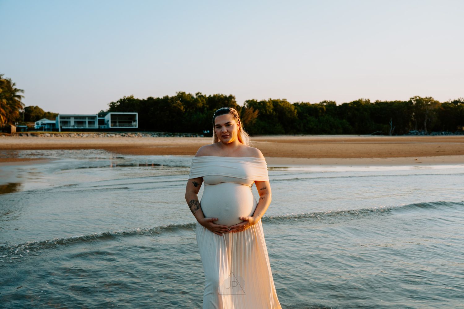 A maternity photo shoot captures a woman in white dress standing in gentle waves at dusk by the shore.