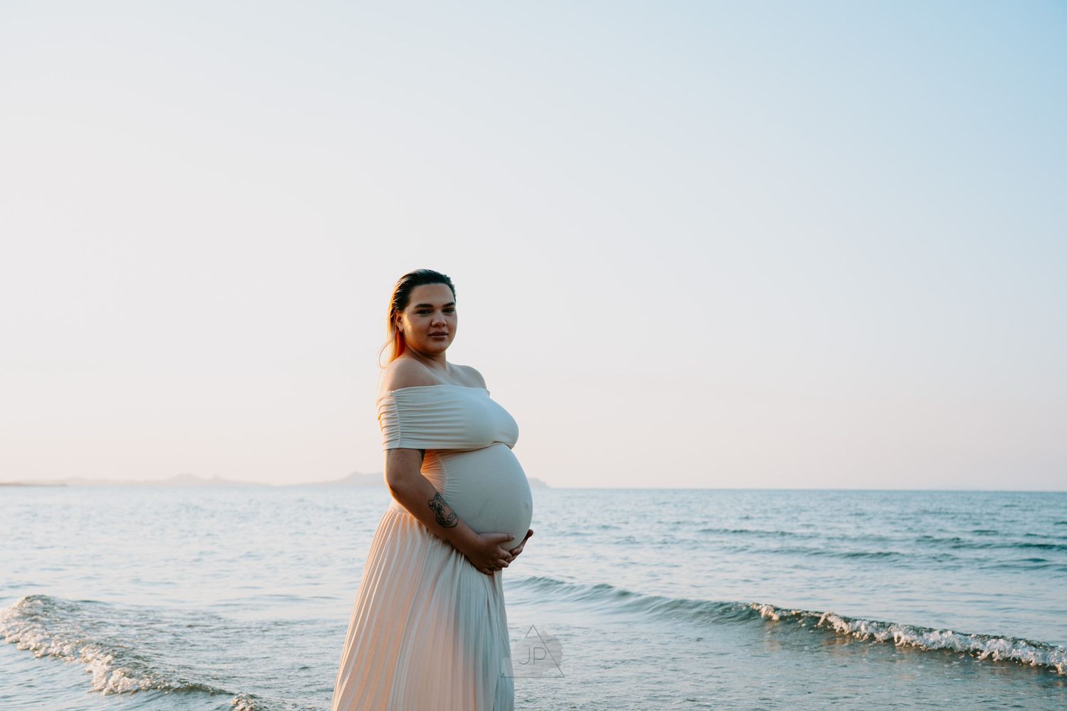 A pregnant woman in a white dress walks along a serene beach at sunset with gentle waves in the background.