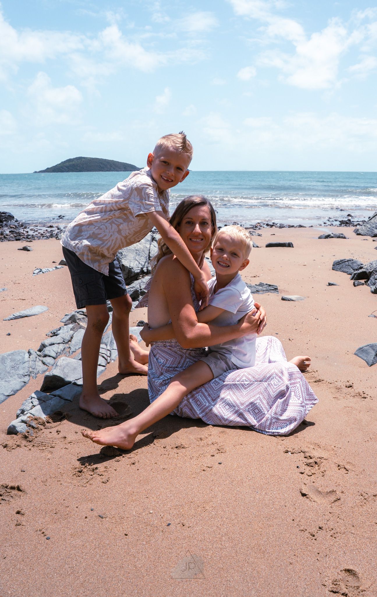 Two people playfully wrestling and laughing together on a sandy beach with waves and rocks in the background.