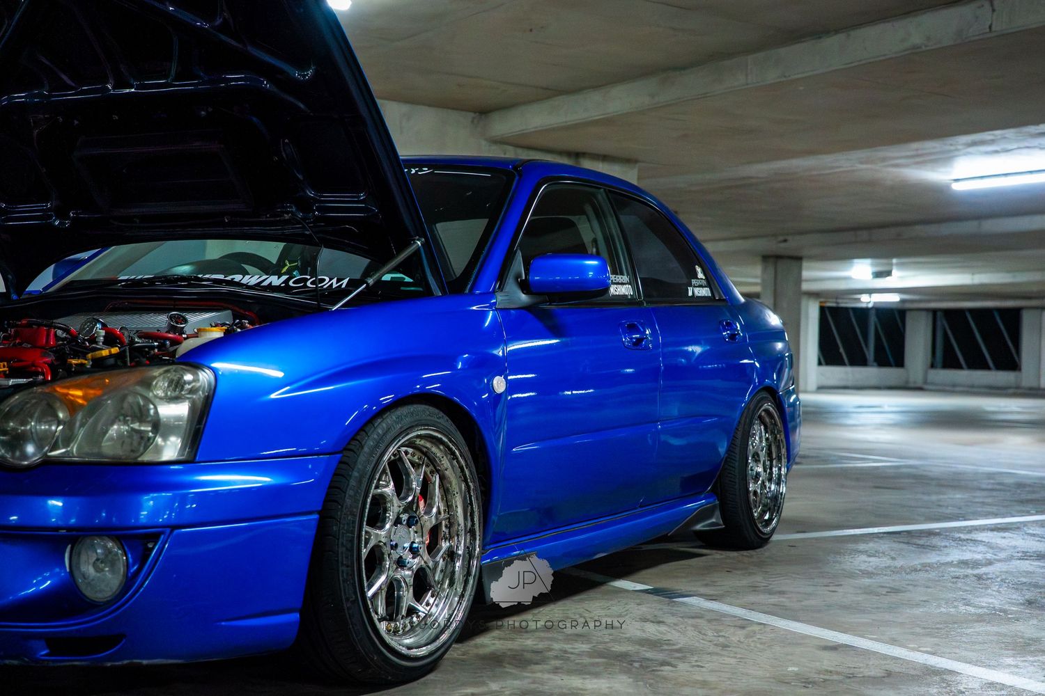 Modified blue Subaru WRX with silver aftermarket wheels sits in a dimly lit parking garage.