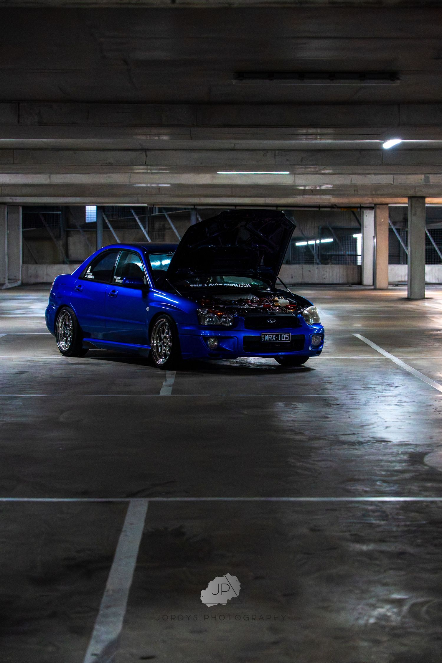 Dark parking garage interior with blue modified sports car visible at multiple angles in parking spaces.