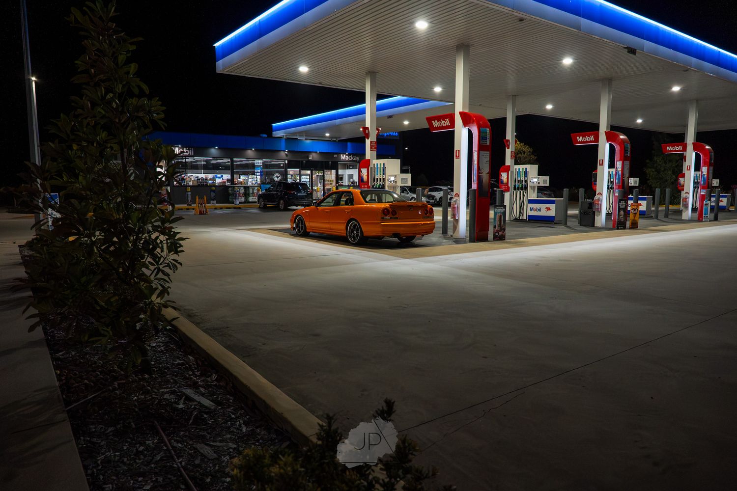 Gas station illuminated at night with bright lights reflecting off parked vehicles and fuel pumps.