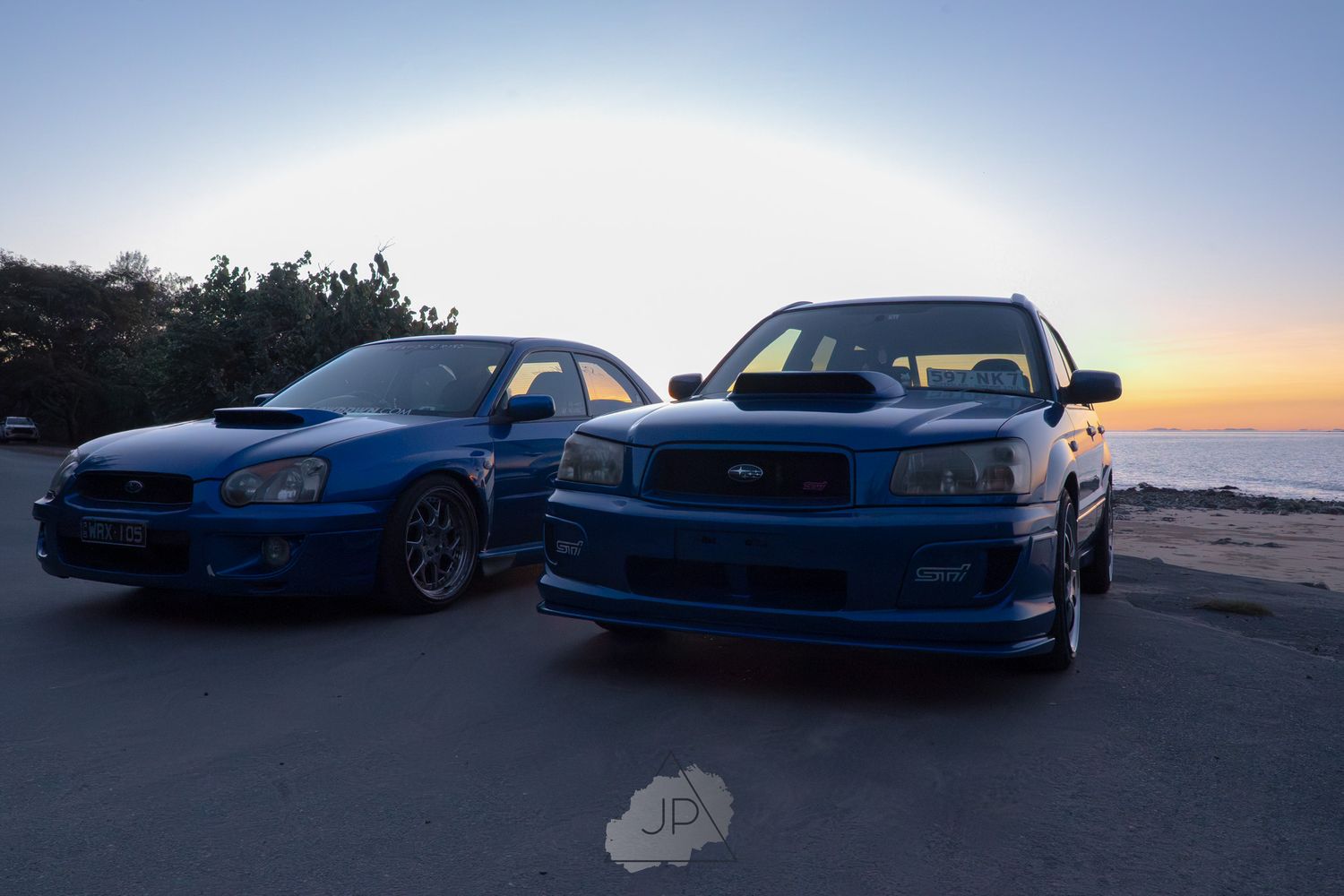 Multiple blue and black sports cars parked along a coastline at sunset create a dramatic silhouette against the sky.