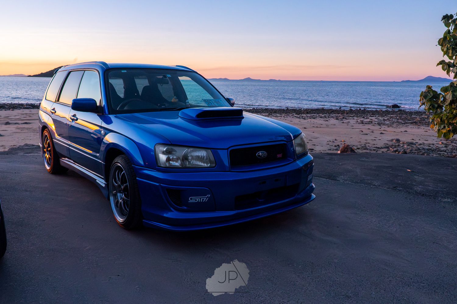 Lowered blue Subaru WRX sits parked at sunset along a coastal road with ocean views.