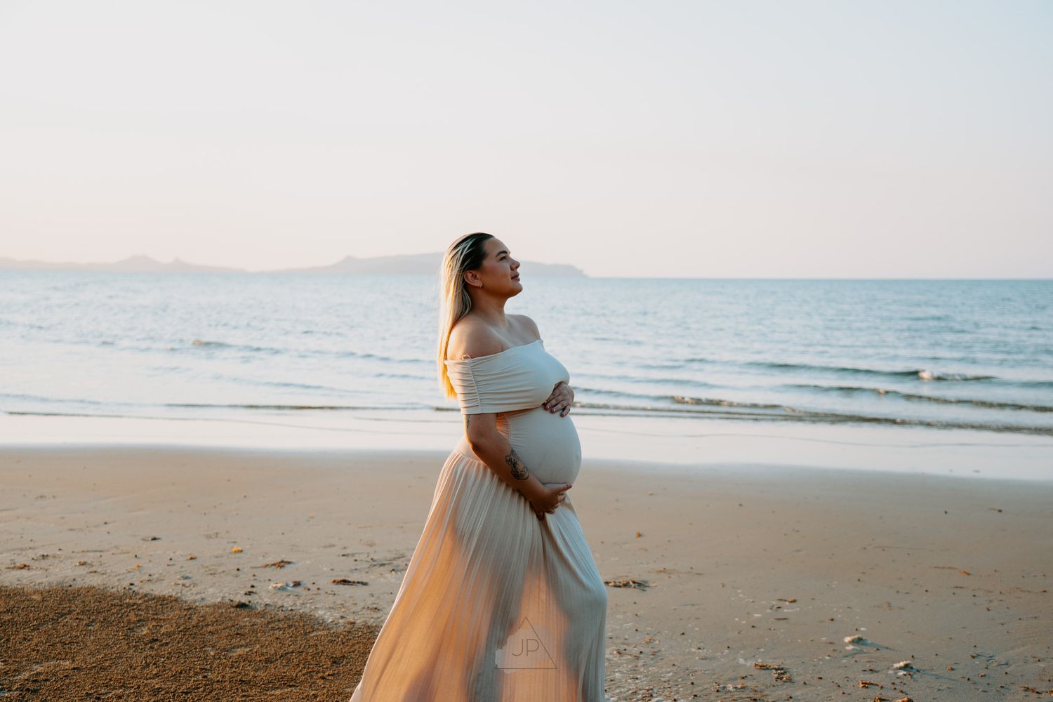 Expectant woman in flowing dress poses on beach at sunset with ocean waves and mountains in background.