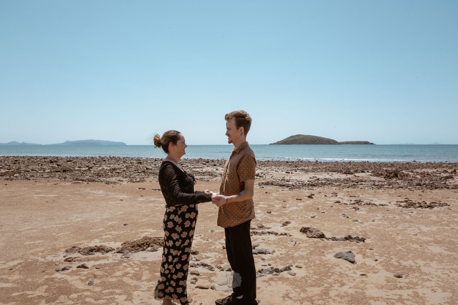 A couple stands together on a scenic beach with mountains in the distance and desert landscape surrounding them.