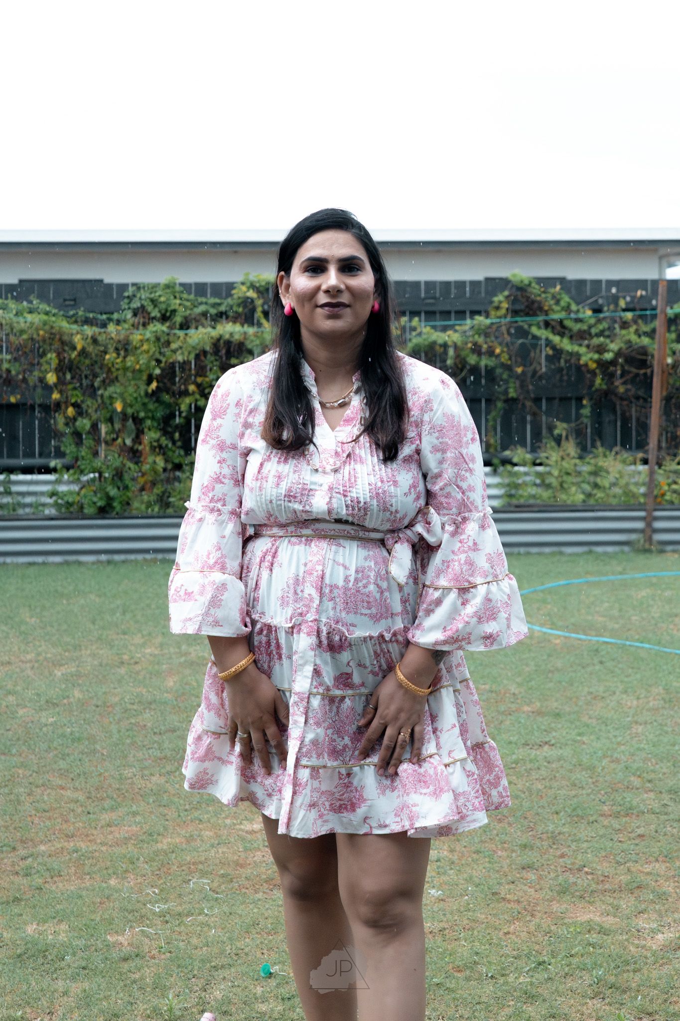 A person wearing a pink floral print dress poses outdoors in a garden setting with green grass and plants in the background.