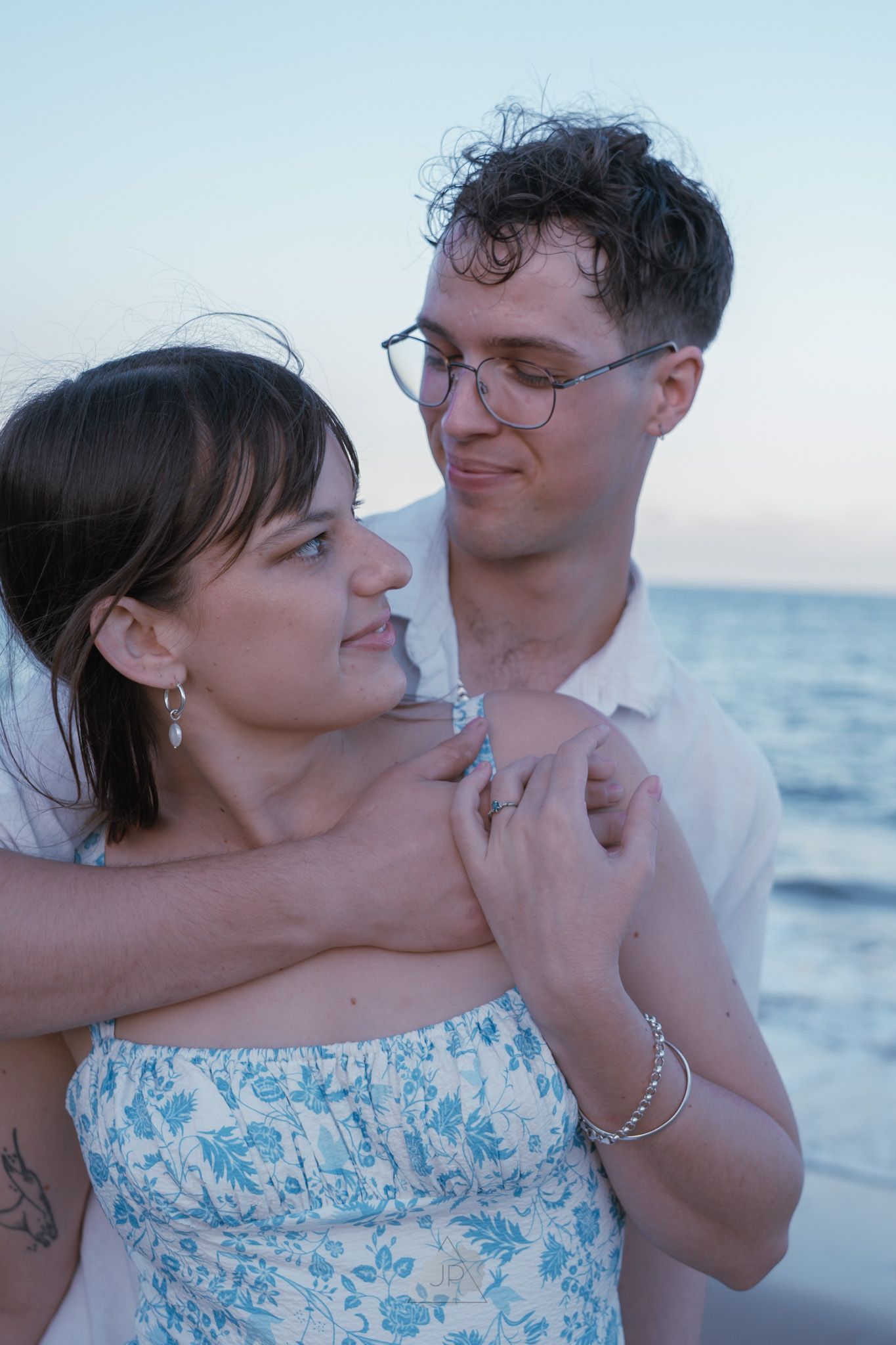A romantic couple embraces on a beach at sunset, with the woman wearing a blue and white floral dress.