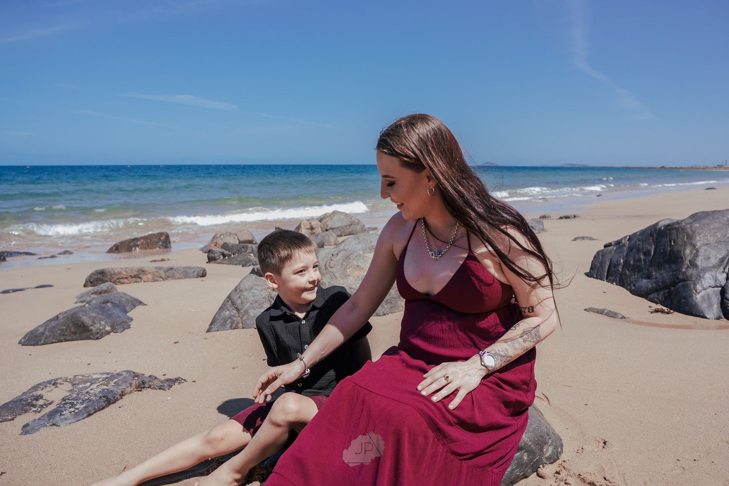 A tender interaction between a child and pregnant person sitting on a sandy beach with large rocks nearby.