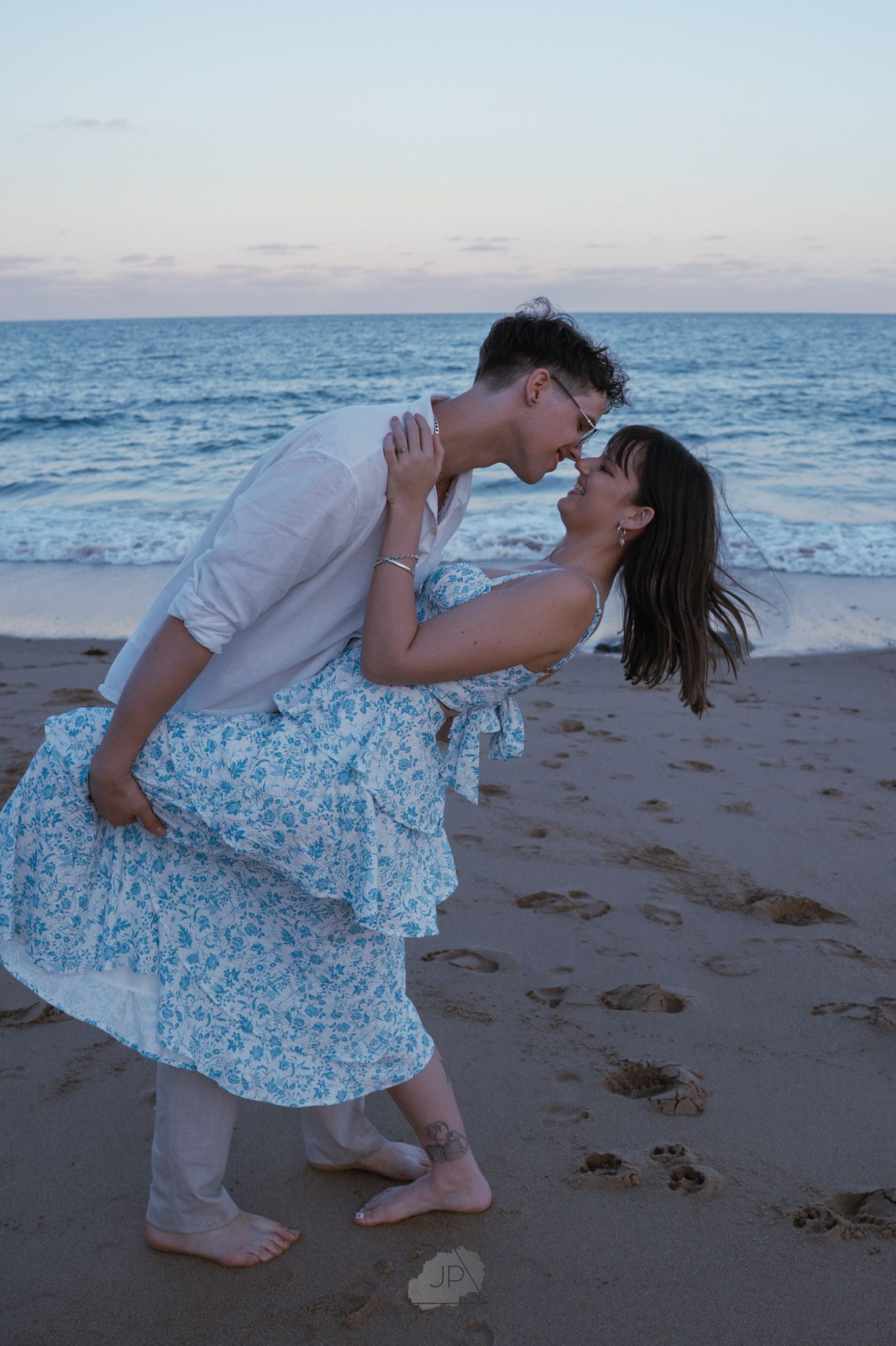 A romantic beach scene at sunset with a couple in an intimate embrace, wearing light blue and white clothing.