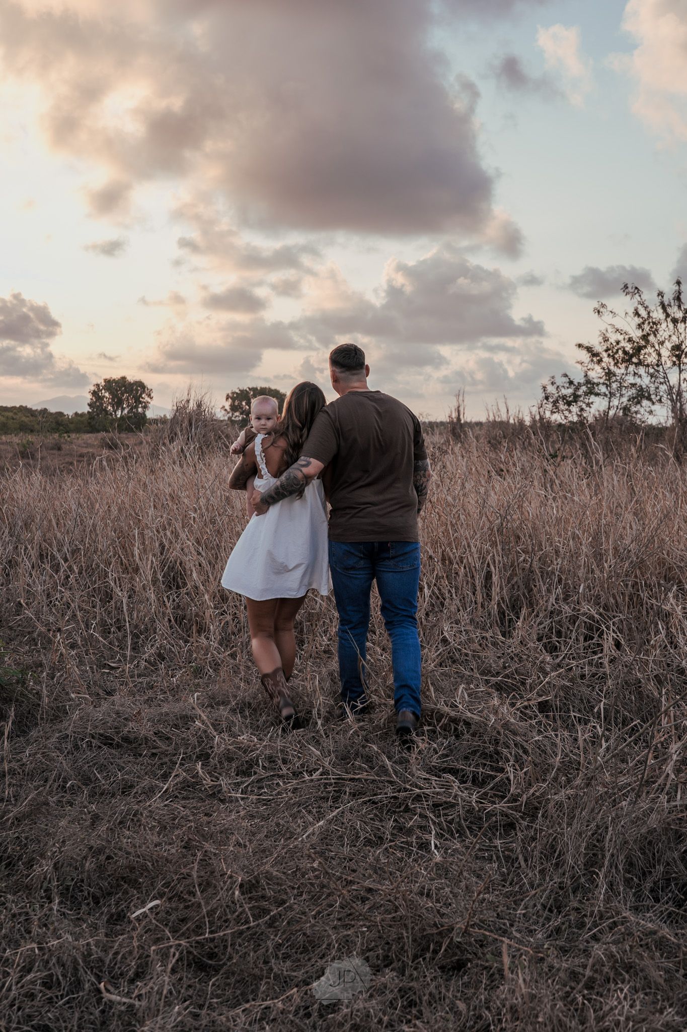 A couple stands together in a rustic field at sunset with dramatic clouds overhead.