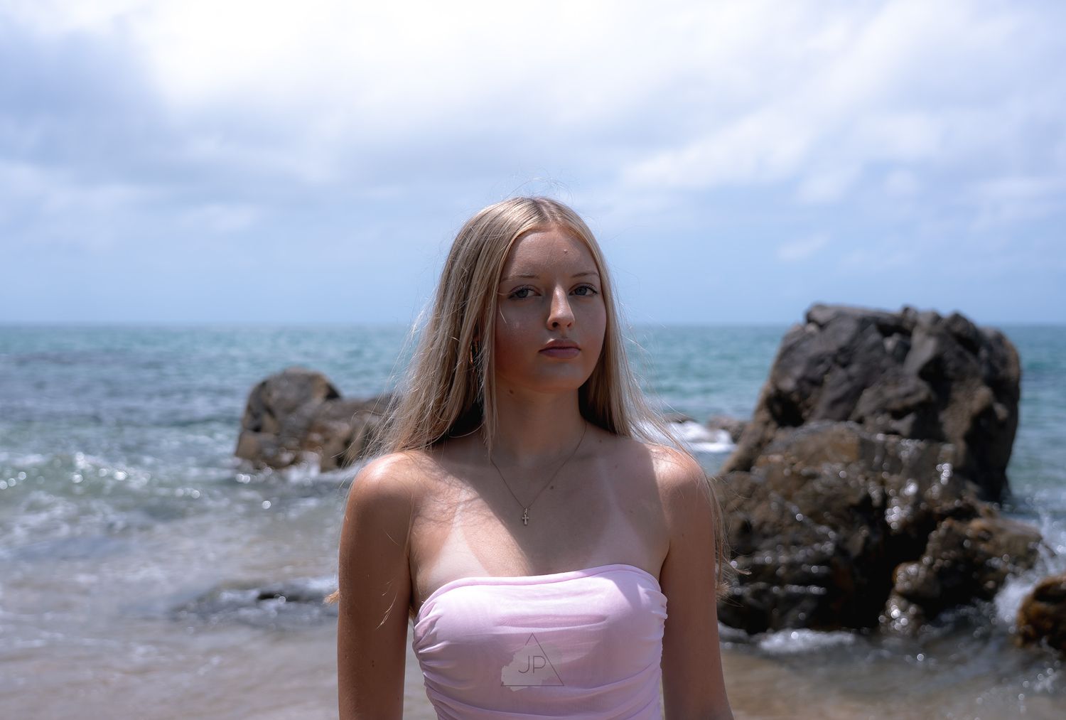 A person in a pink swimsuit stands near rocky coastal waters on a sunny day.