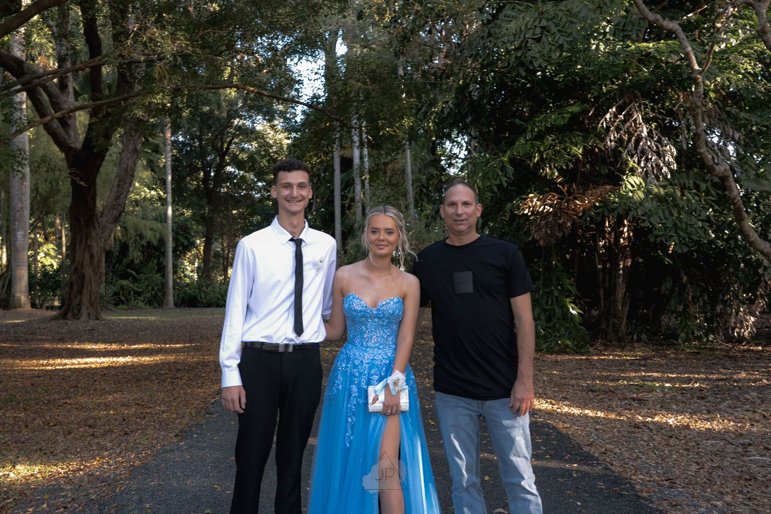 A group poses for outdoor photos in formal attire under tree-lined paths on a sunny autumn day.