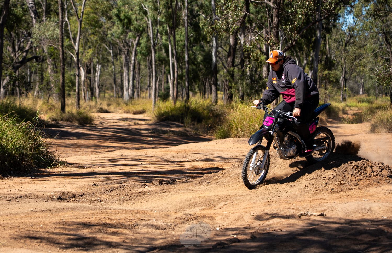 A dirt bike rider navigates a winding trail through an Australian bushland setting with scattered gum trees.
