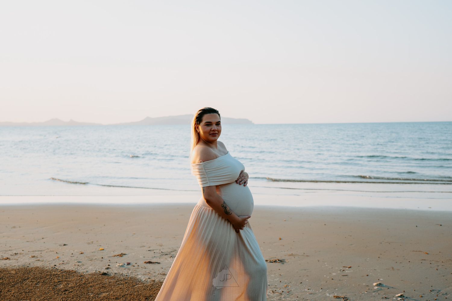 Pregnant woman in flowing white dress walking along peaceful beach at sunset with mountains in background.