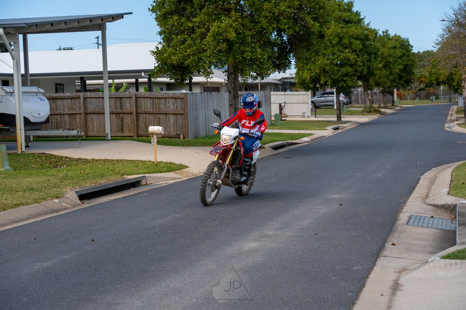 A motorcyclist rides a red dirt bike down a residential street on a sunny day.