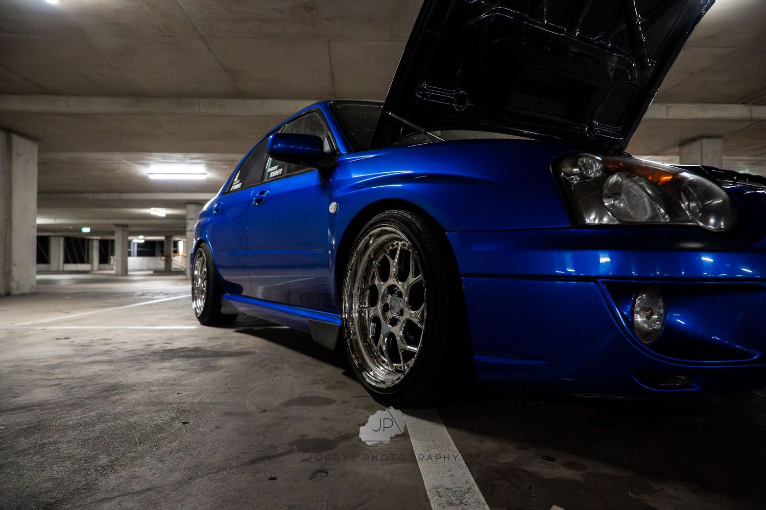Multiple blue sports cars parked in a dimly lit concrete parking garage with stylish aftermarket wheels.
