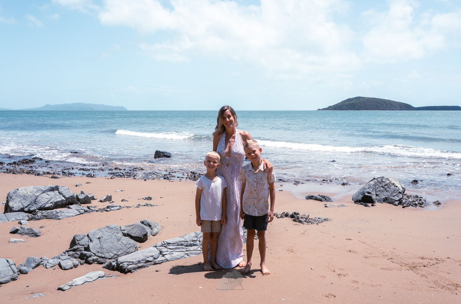 People walking on a scenic beach with rocky outcrops and blue ocean waves under a bright sky.