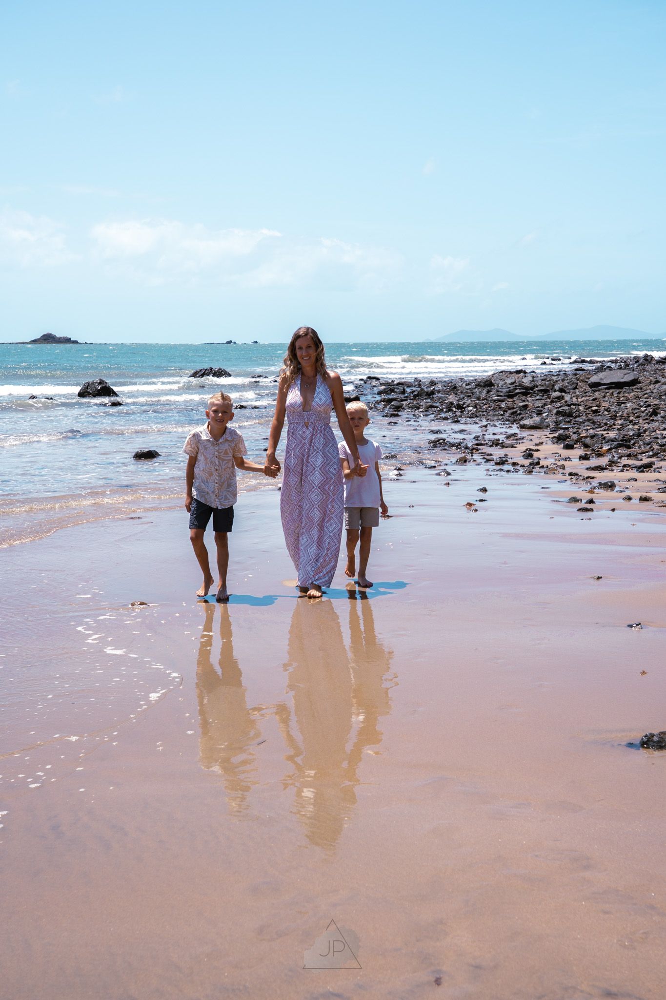 Three people walk along a scenic pink sandy beach with rocky outcrops and blue ocean waves in the distance.