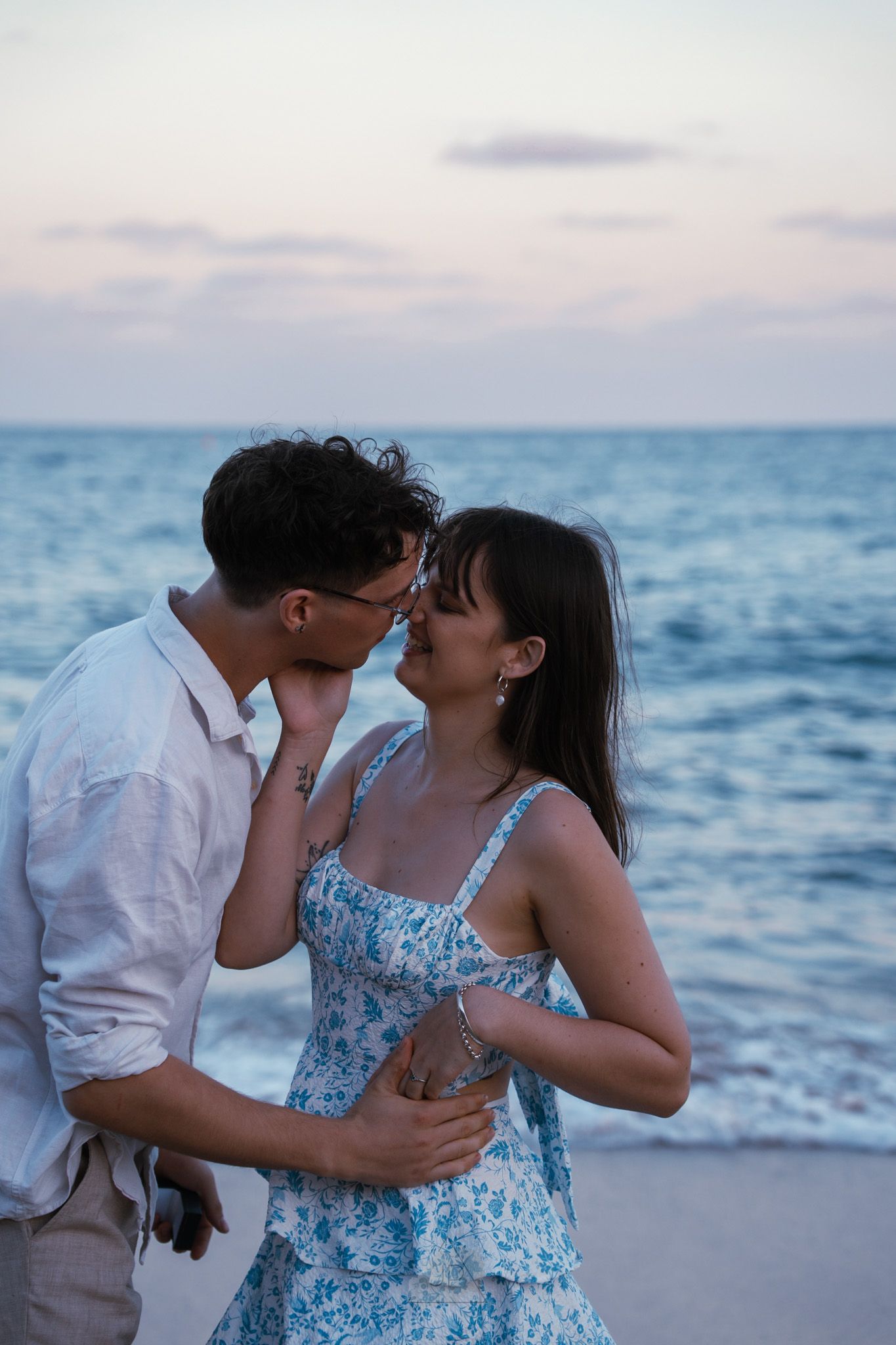 A romantic couple shares tender moments on a beach at sunset with ocean waves in the background.
