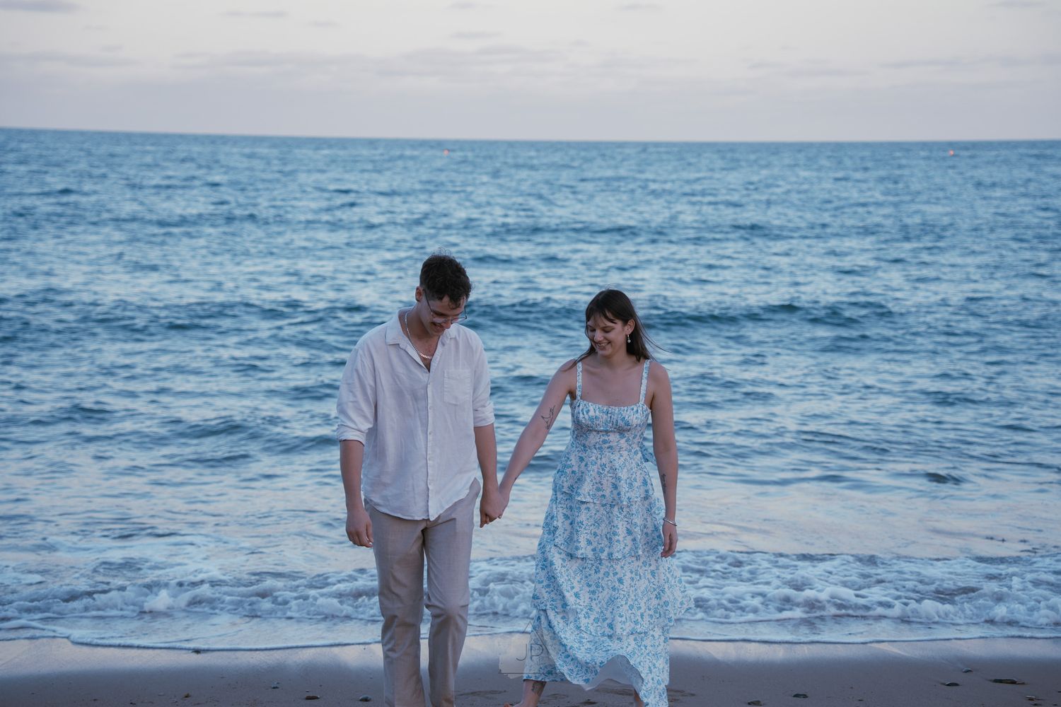 A couple walks hand in hand along a scenic beach with blue ocean waves crashing behind them.