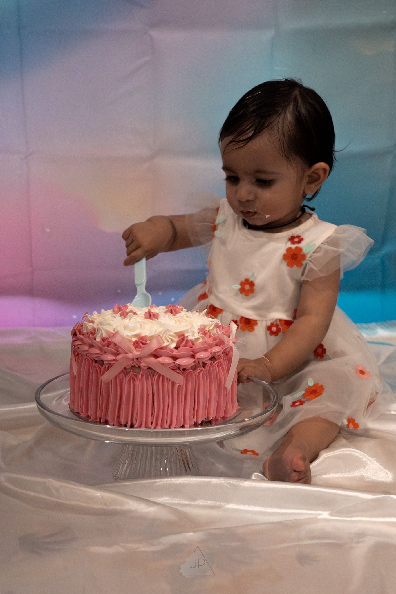 A pink frosted cake with floral decorations is displayed on a clear glass pedestal stand against a dreamy background.
