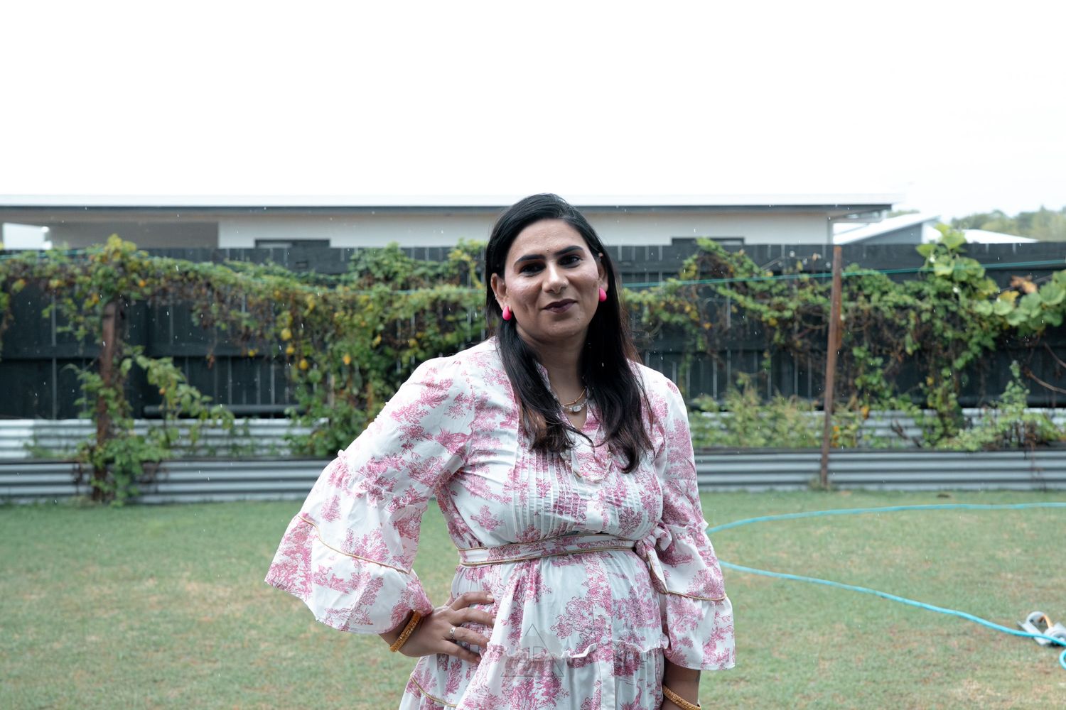 Person in a pink floral dress with flared sleeves poses in a garden with greenery in the background.
