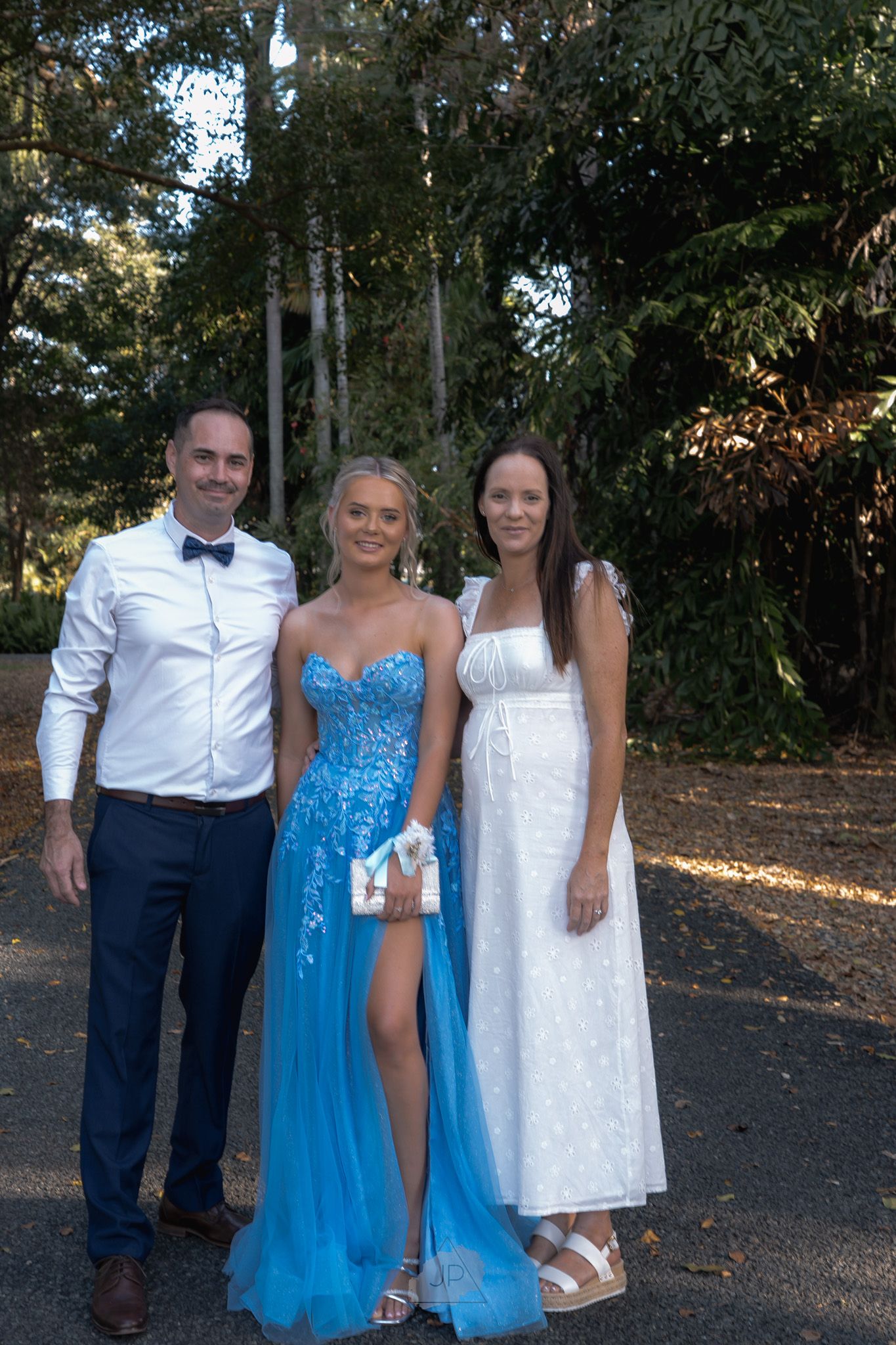 Three people pose together outdoors wearing formal attire including a white dress shirt and bright blue dress.