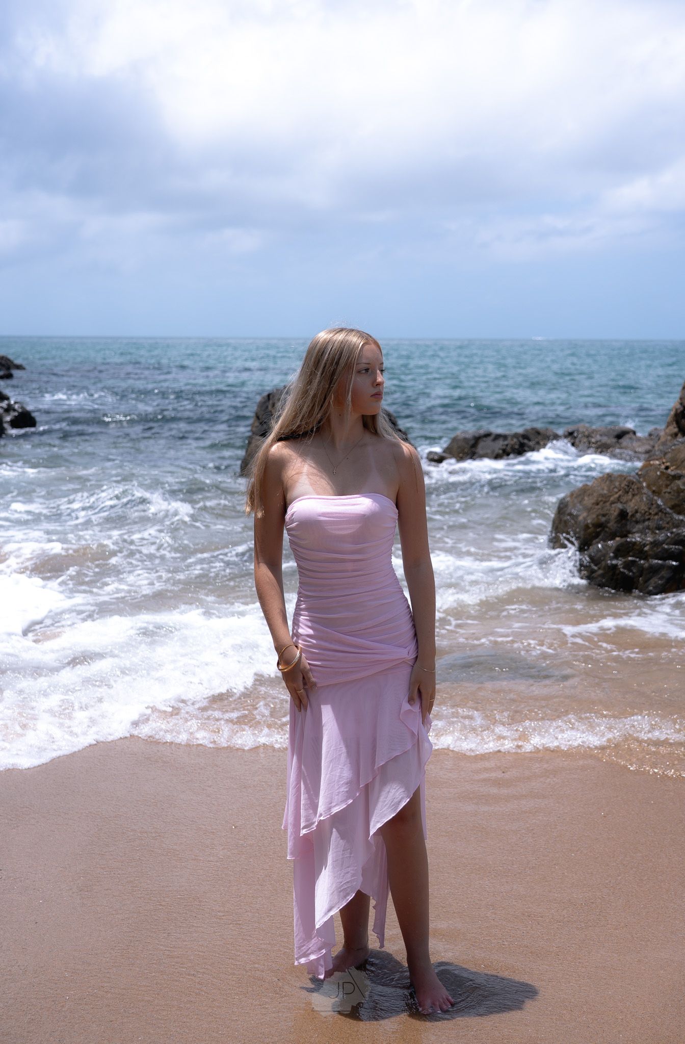 A person wearing a pink dress stands on a sandy beach with ocean waves and rocks in the background.