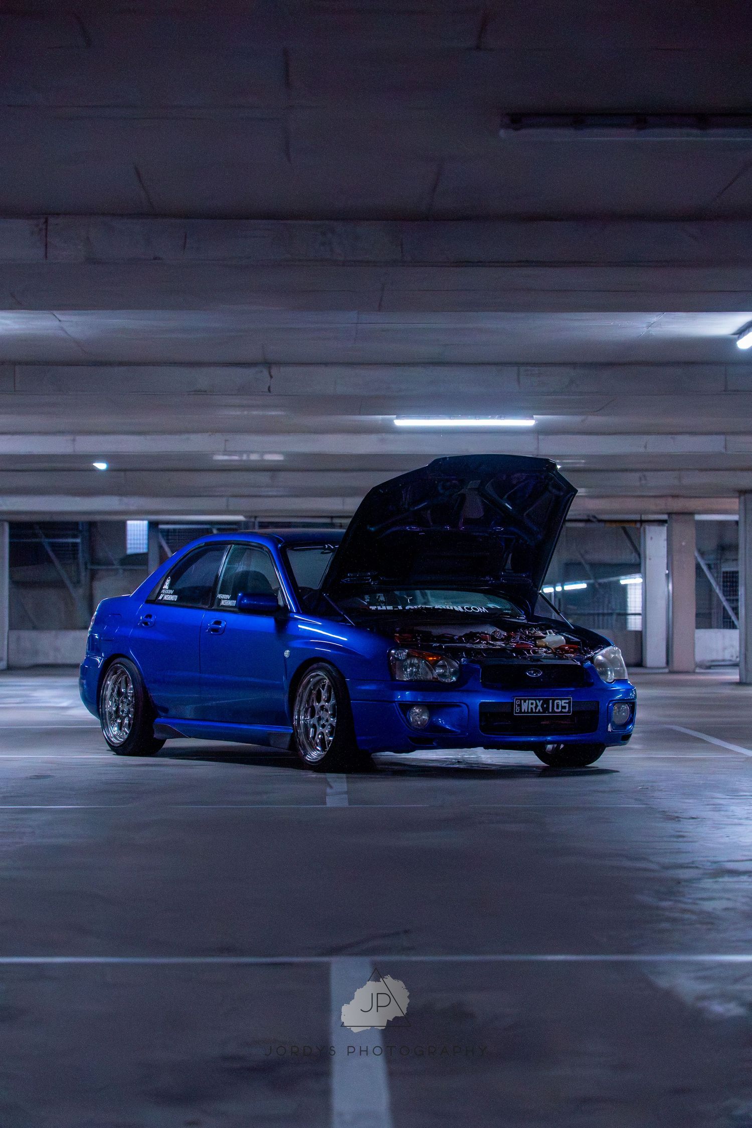 Low angle view of a modified blue sports car parked in a dimly lit parking garage showing its sleek profile.